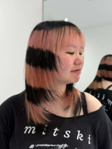 A young woman at Assort London showcasing a creative 'Angel Halo' hair color inspired by Alysa Liu. The hairstyle features a medium-length, layered Japanese cut with a peach-pink base and two distinct, horizontal jet-black bands (rings) circling the hair. The look includes soft wispy bangs and face-framing layers, highlighting the precision of the horizontal 'ring dye' technique.